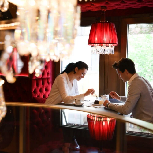 A couple sits in a cozy window alcove with deep red, quilted upholstered walls and red crystal lamps.