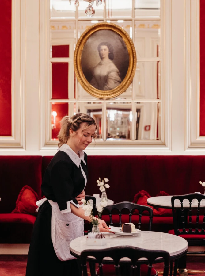 Staff member at Café Sacher Wien serving dessert at marble table with red velvet seating and classic décor