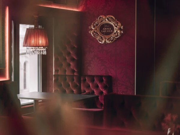 In the center of the picture hangs an artistically framed emblem bearing the inscription “Salon Anna Sacher”. To the left, natural light streams in through a window, below it hangs a classic lamp with crystal pendants, casting a warm glow.