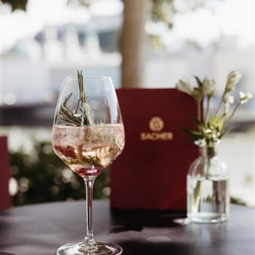 Aperitif in a wine glass with a sprig of rosemary on a terrace table, with a Sacher menu and a small vase with flowers in the background