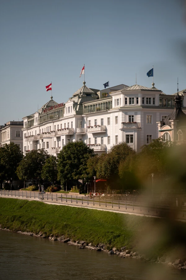 Hotel Sacher Salzburg along the Salzach River with flags waving above, surrounded by trees and a riverside promenade