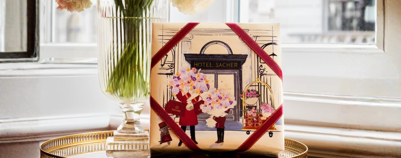 Gift box of the Original Sacher-Torte Mother’s Day edition with a red ribbon and illustration placed by a window, next to a vase with flowers