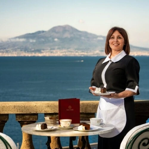 Sacher service team member serving Original Sacher-Torte on a terrace with sea view and Mount Vesuvius in the background.