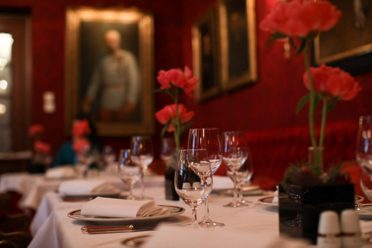 An elegant dining room at Hotel Sacher, adorned with red walls, classic portraits, and tables set with fine glassware and blooming pink flowers.