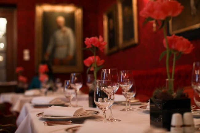 An elegant dining room at Hotel Sacher, adorned with red walls, classic portraits, and tables set with fine glassware and blooming pink flowers.