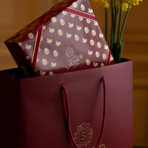 Original Sacher-Torte gift box in a Sacher shopping bag at Hotel Sacher Vienna with yellow flowers and elegant interior backdrop.