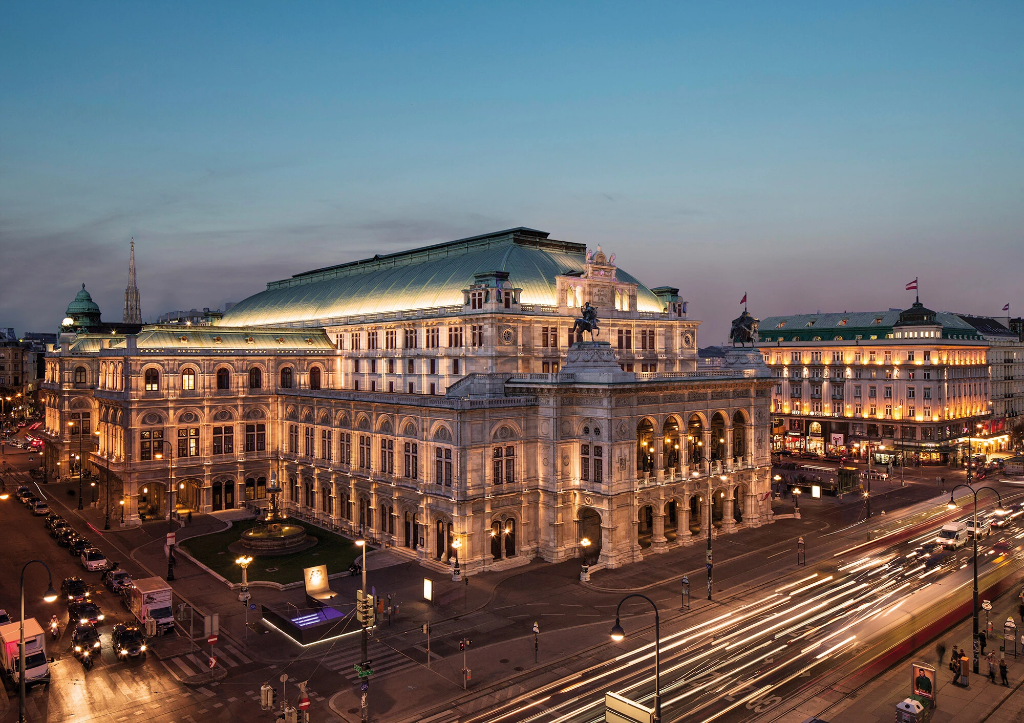 The Vienna State Opera glows majestically at dusk, with the elegant Hotel Sacher illuminated just across the bustling Ringstraße.
