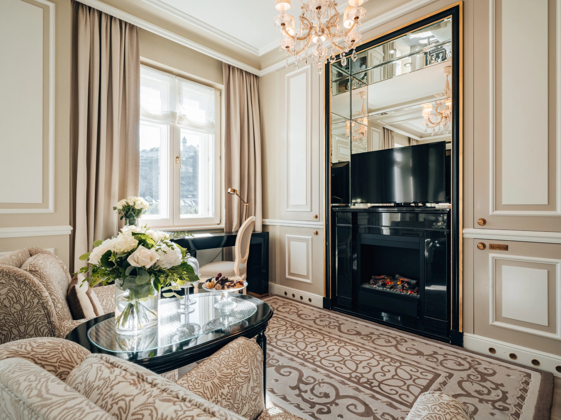 Seating area in the Deluxe Room with View at Hotel Sacher Salzburg, featuring a fireplace, elegant writing desk, large windows and classic details