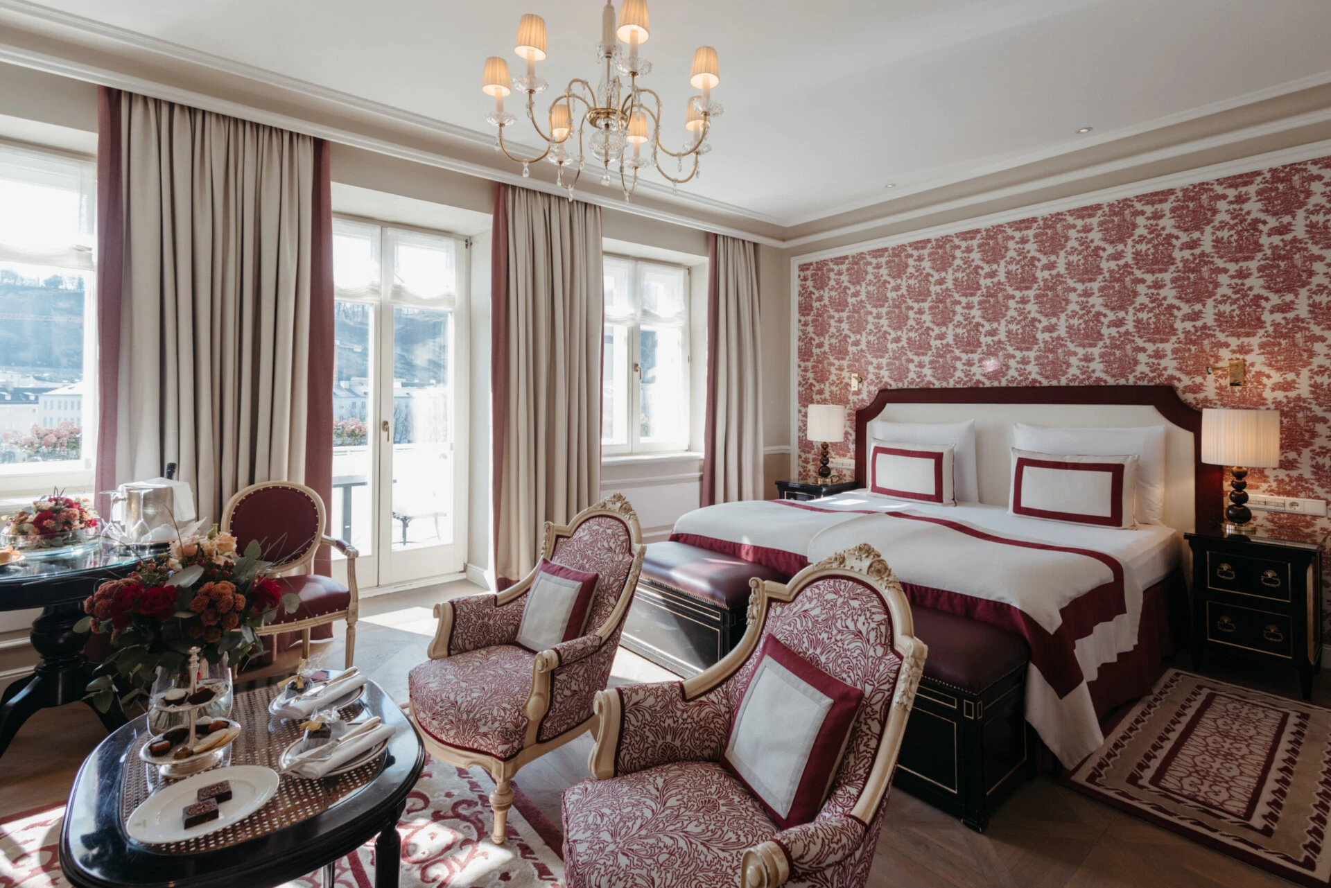 Junior Suite with View at Hotel Sacher Salzburg, featuring an elegant double bed, classic armchairs, red patterned details and large windows