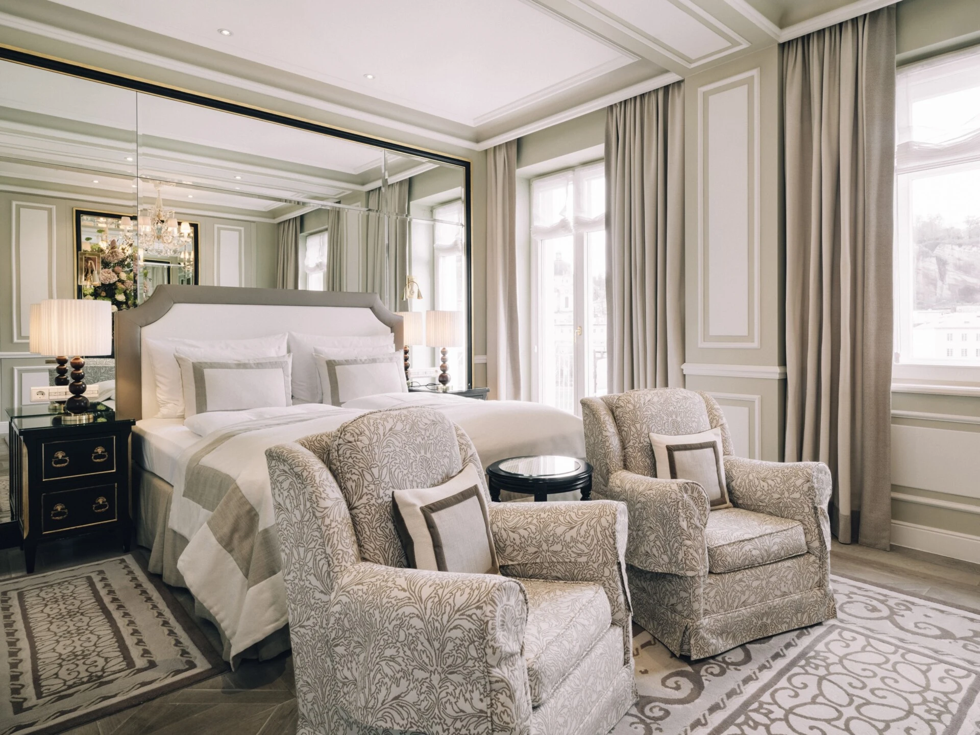 Junior Suite with View at Hotel Sacher Salzburg, featuring an elegant bed, patterned armchairs, classic interiors and large windows overlooking Salzburg's Old Town