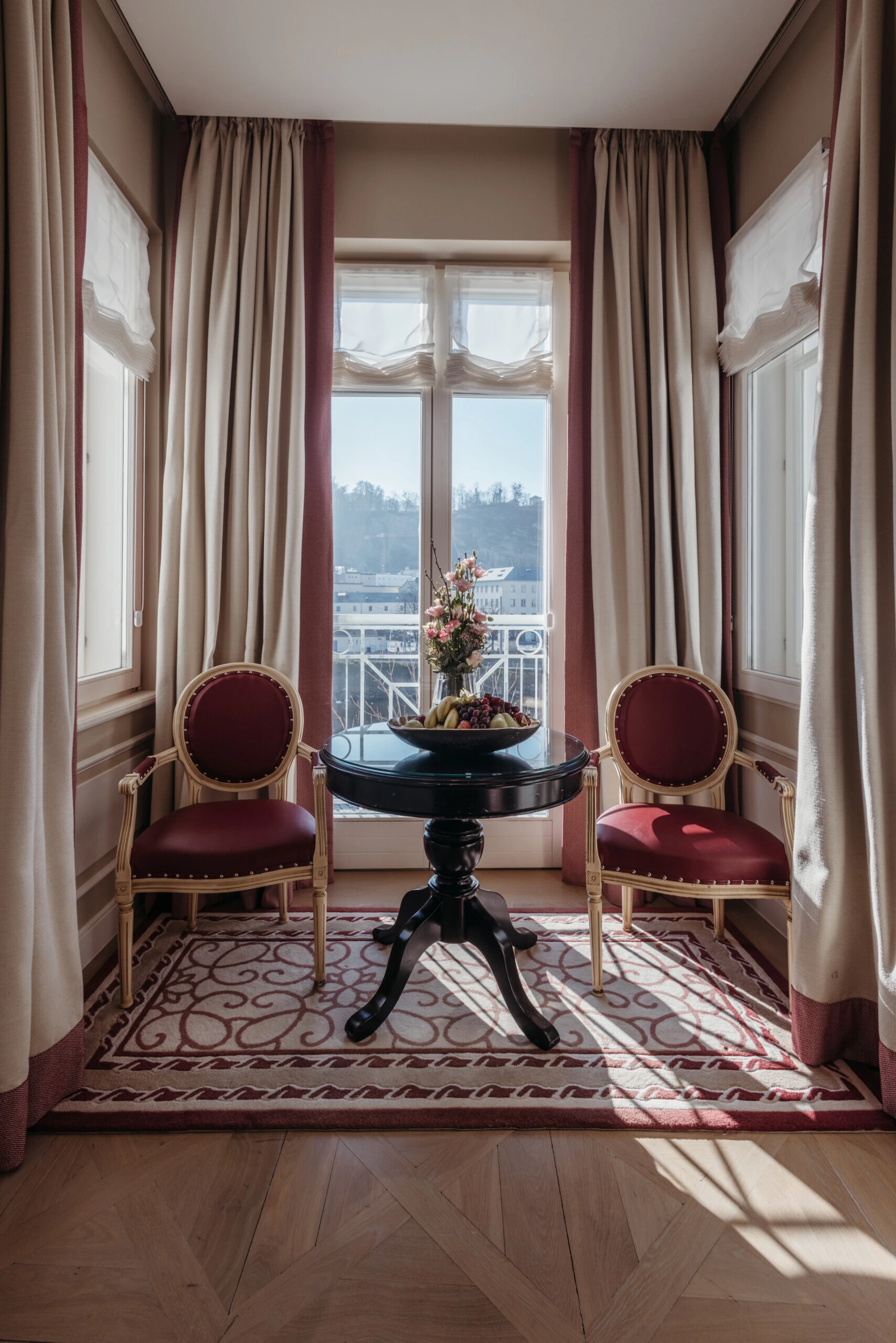 Seating area in the Family Suite with a view at the Hotel Sacher in Salzburg, featuring a round table, red chairs, and large windows