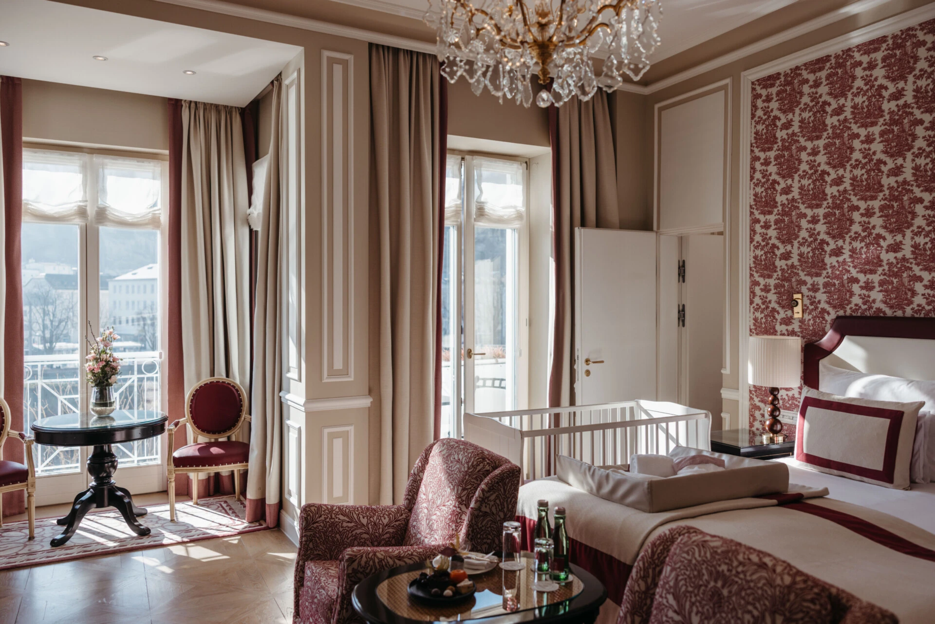 Family suite with a view at the Hotel Sacher Salzburg, featuring a bedroom, sitting area, crib, and elegant red patterned accents