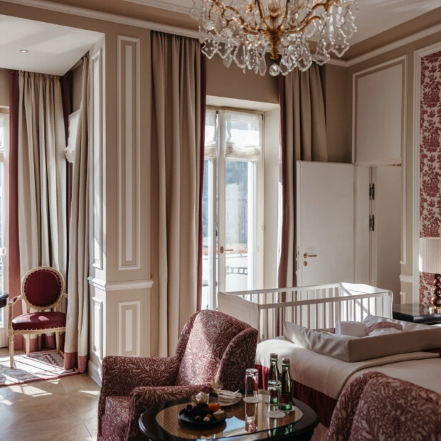 Family suite with a view at the Hotel Sacher Salzburg, featuring a bedroom, sitting area, crib, and elegant red patterned accents