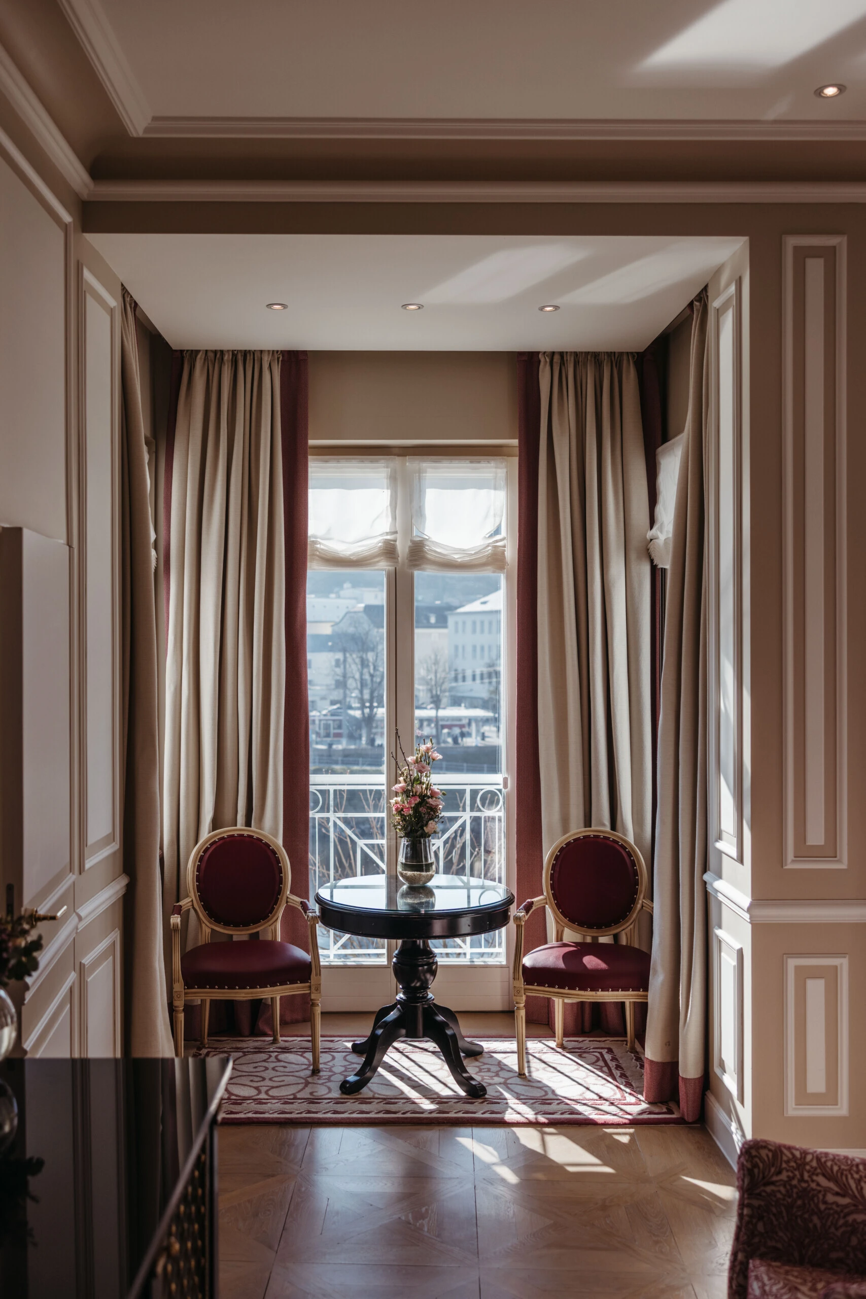 Seating area in the Family Suite with a view at the Hotel Sacher in Salzburg, featuring a round table, red chairs, and large windows