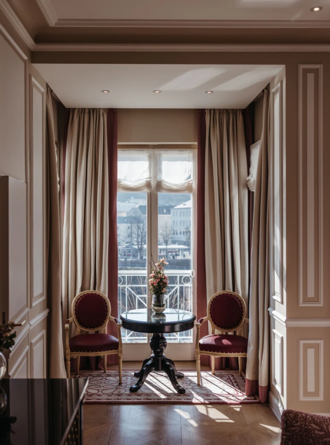 Seating area in the Family Suite with a view at the Hotel Sacher in Salzburg, featuring a round table, red chairs, and large windows
