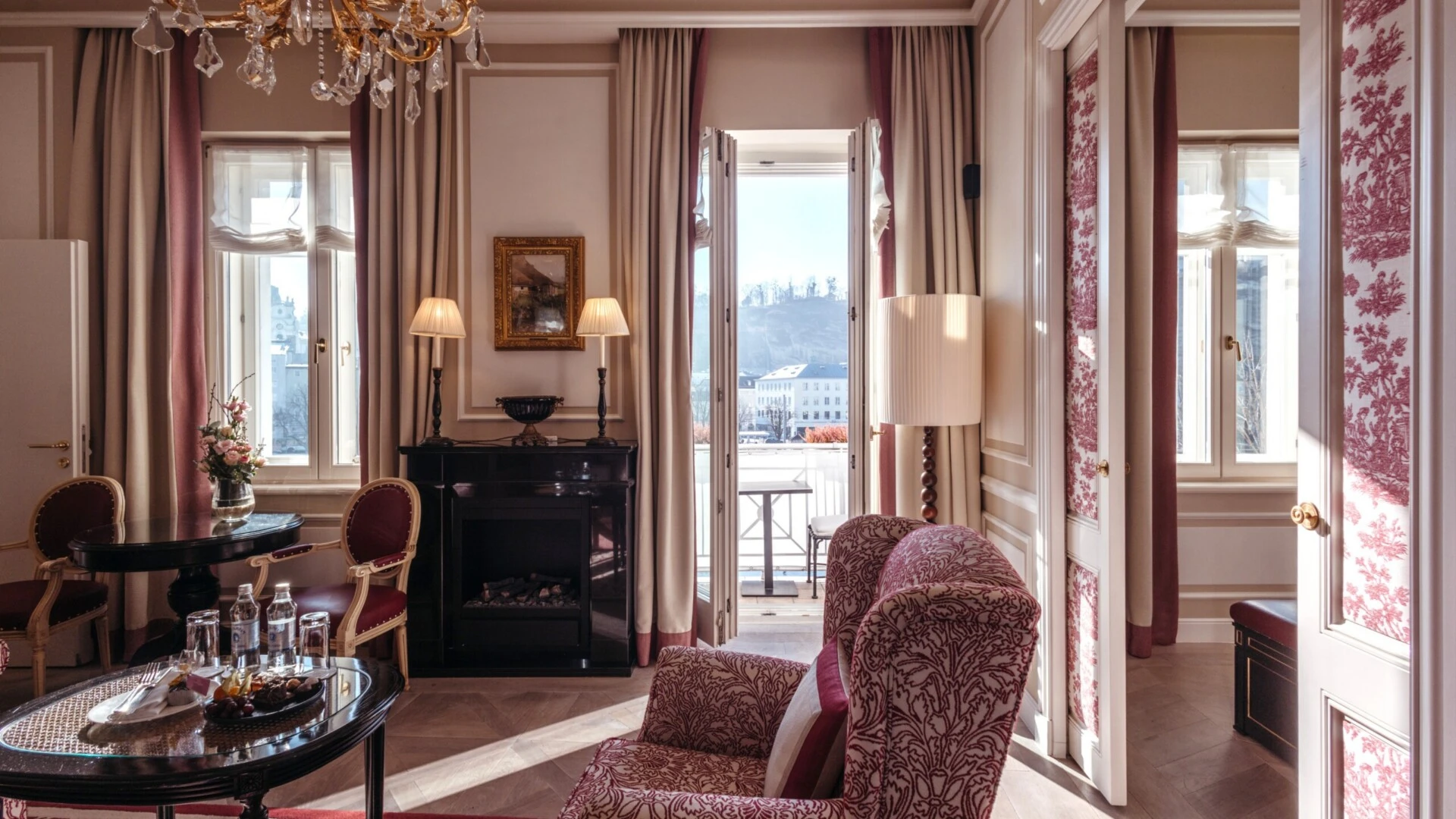 Family suite with a view at the Hotel Sacher in Salzburg, featuring a bedroom, a sitting area, and elegant red patterned accents