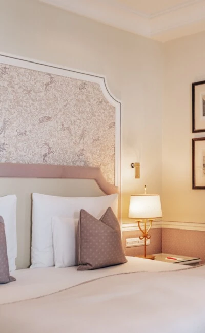 Close-up of the Junior Suite at Hotel Sacher Salzburg featuring an elegant bed, decorative headboard, classic lighting and stylish interiors in soft rose tones.