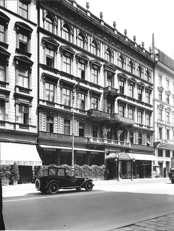 A black-and-white photo of the historic Hotel Sacher building, showcasing its grand façade and vintage cars parked along the street.