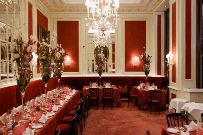 Festively set dining area at Café Sacher Vienna with red velvet banquettes, crystal chandeliers and tall floral arrangements.