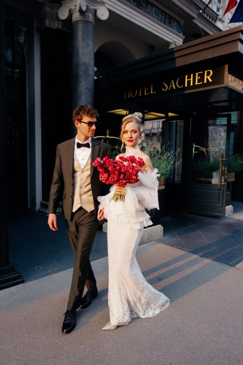 A bride and groom in front of the entrance to the Hotel Sacher Vienna, with the bride holding a red bouquet.