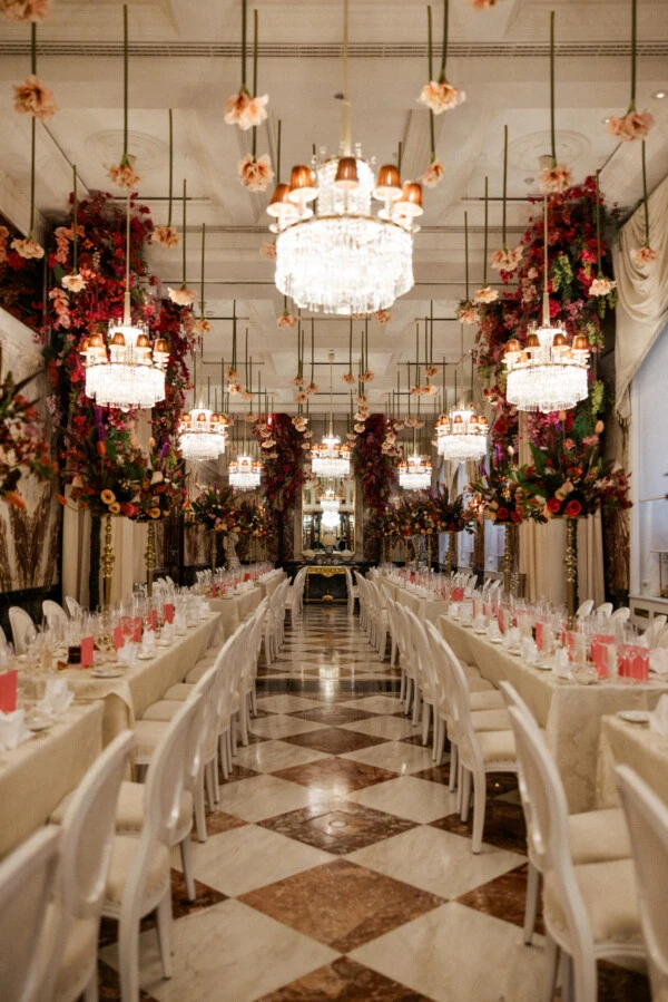 Elegant banquet hall with long dining tables, crystal chandeliers and abundant red and pink floral arrangements.