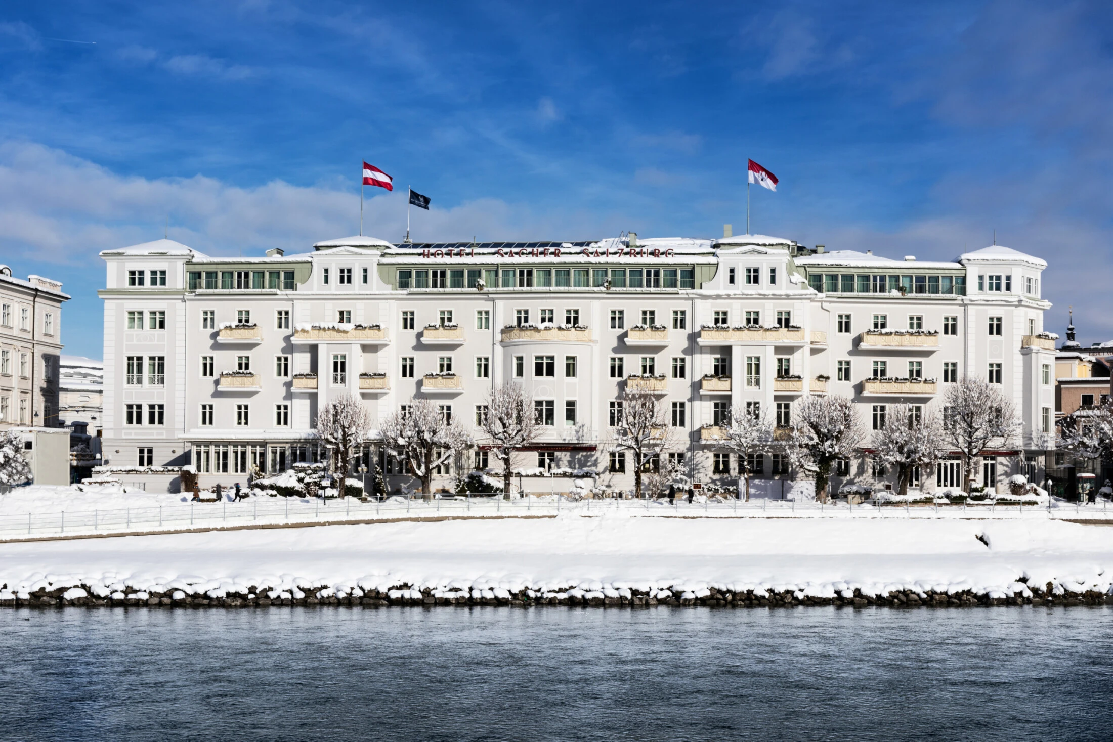 Exterior view of Hotel Sacher Salzburg in winter, with its snow-covered façade and the Salzach River in the foreground.