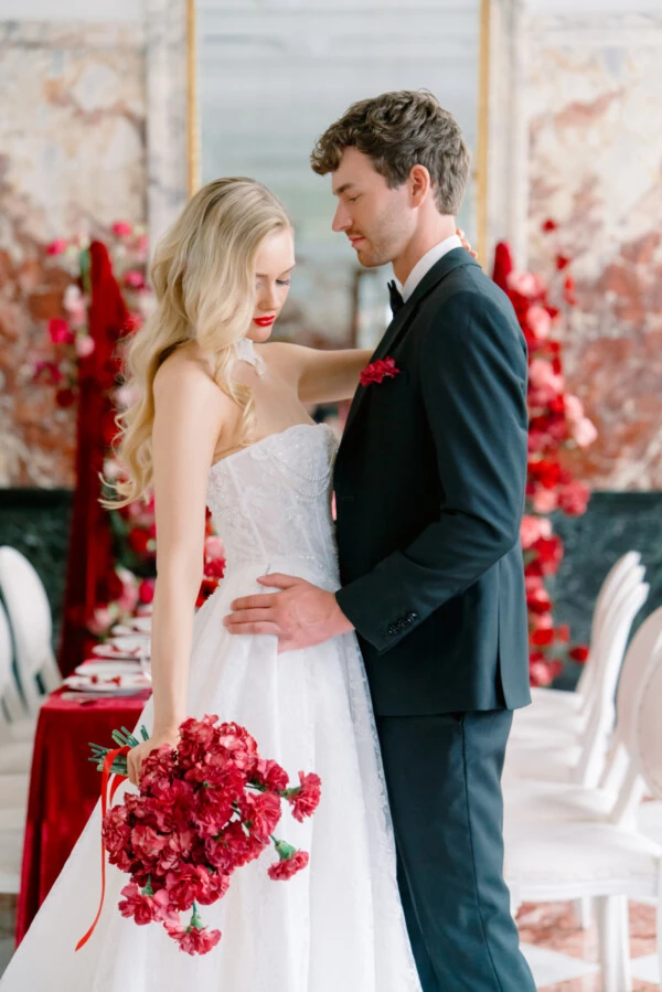 Bride and groom in formal wedding attire before red floral décor, with the bride holding a lush red bouquet.