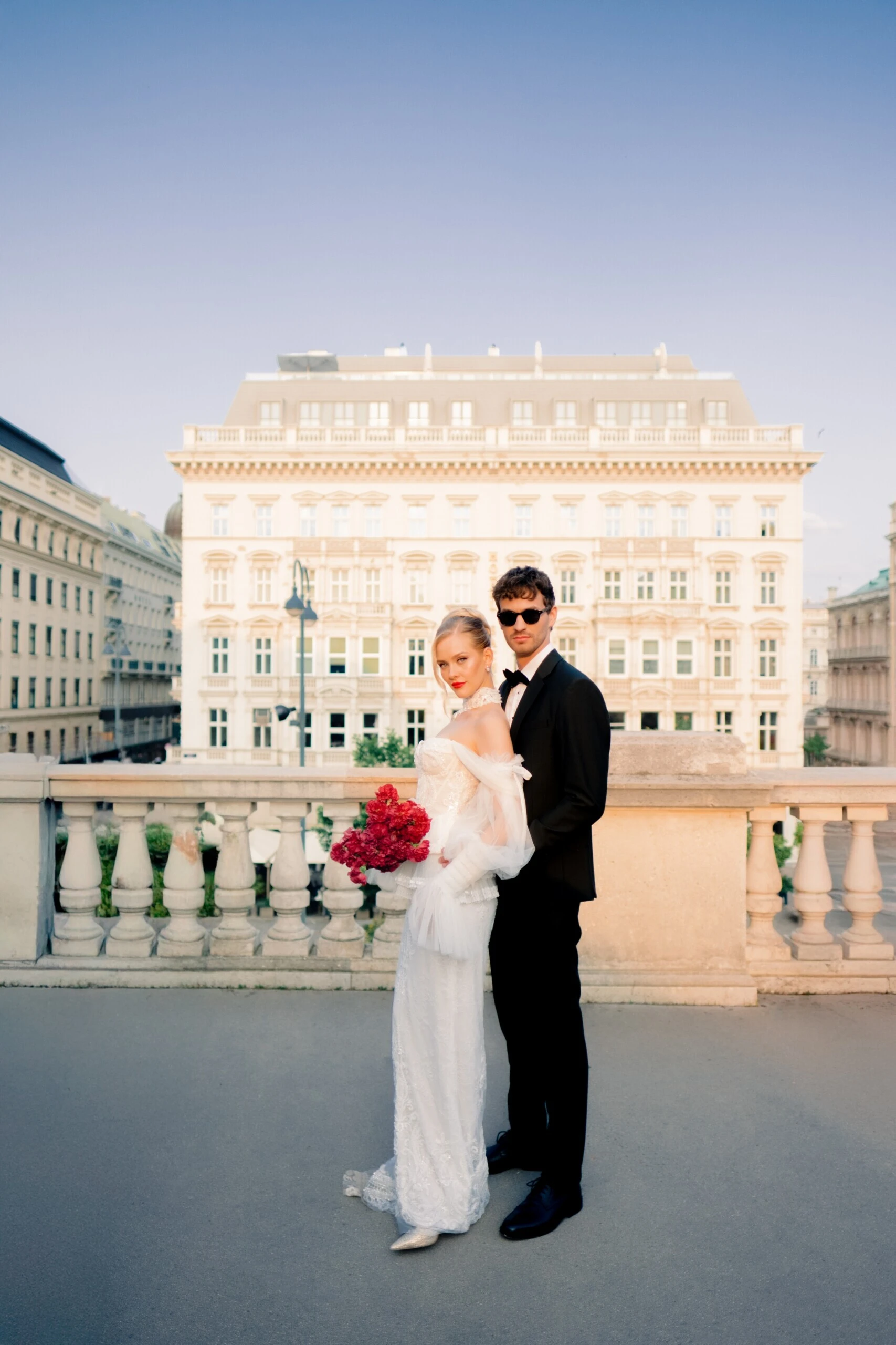 Bride and groom pose with a red bouquet on an elegant terrace against historic city architecture.