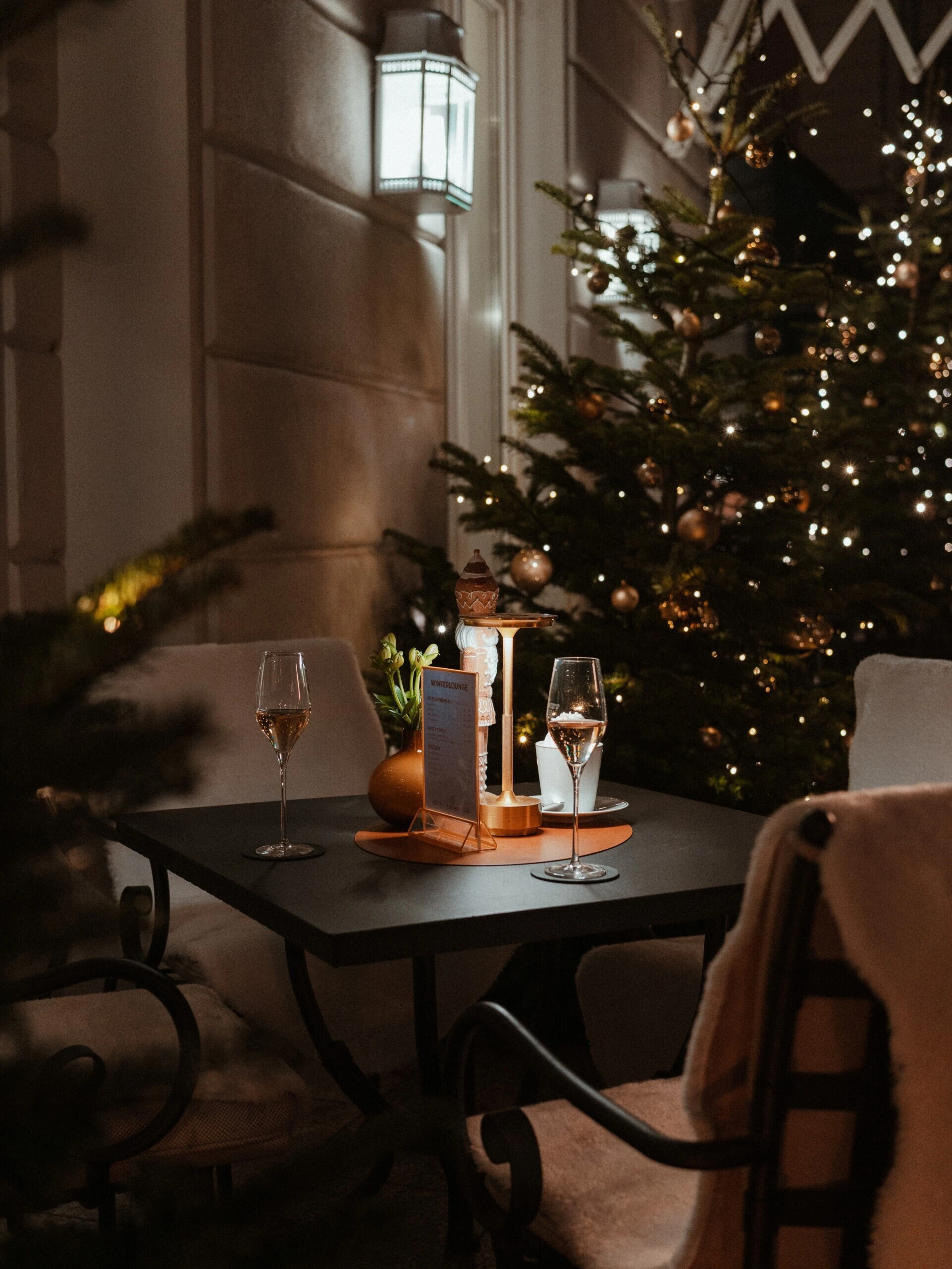 Luxury outdoor winter patio setting with two champagne flutes, a Christmas tree lit with warm bokeh lights, and a nutcracker figurine on a small table.