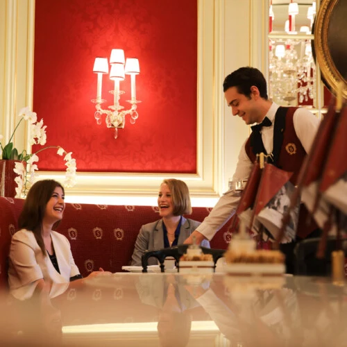 A warm moment at Café Sacher Wien as a smiling waiter serves two guests seated in the elegant red-upholstered interior beneath classic chandeliers and portraits.