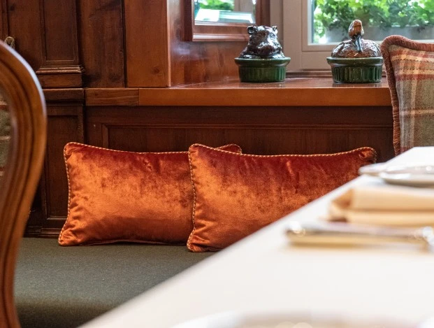 A cozy restaurant corner with orange velvet pillows, plaid cushions, and ceramic animal decorations on the windowsill.