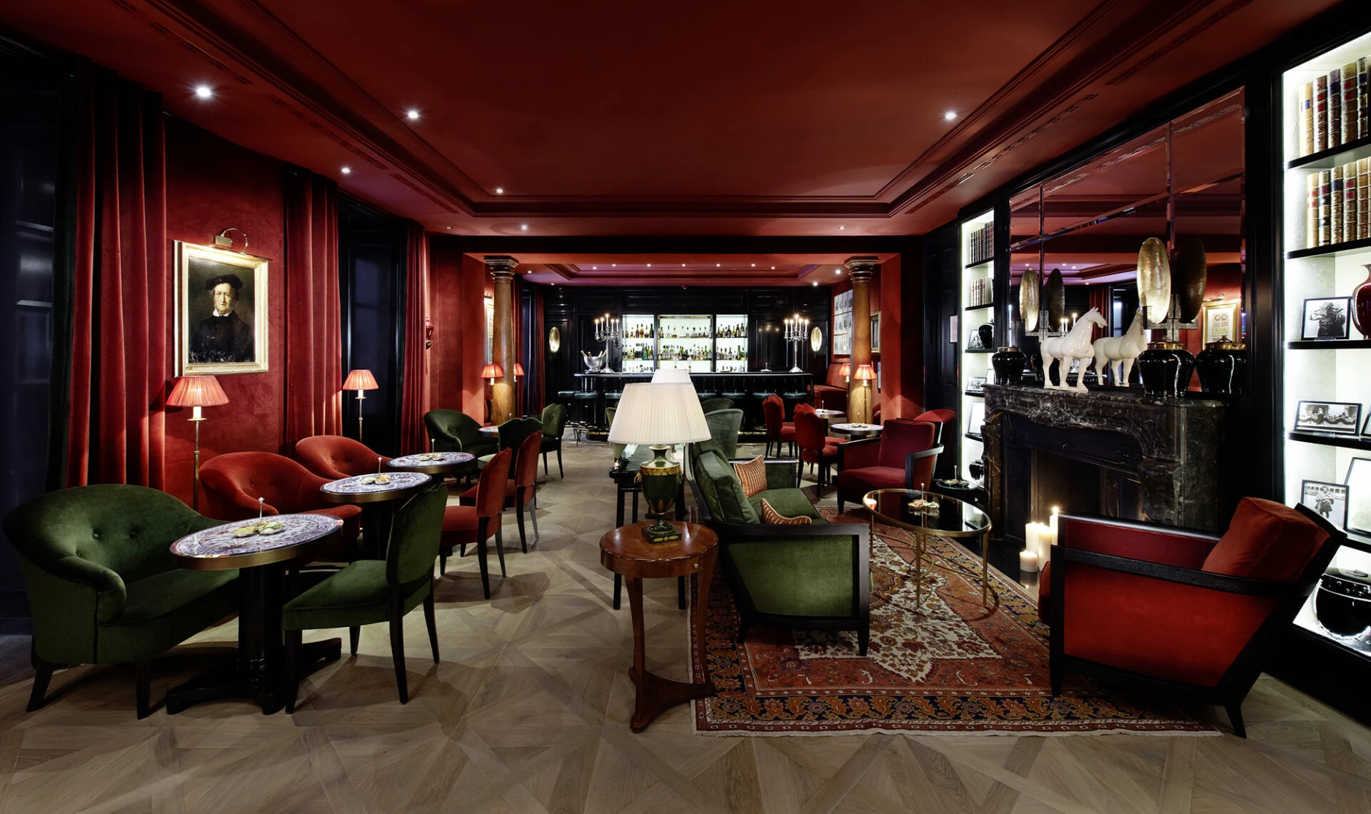 An elegant hotel bar lounge features plush red and green velvet seating, a marble fireplace, and a warmly lit ambiance perfect for refined relaxation.