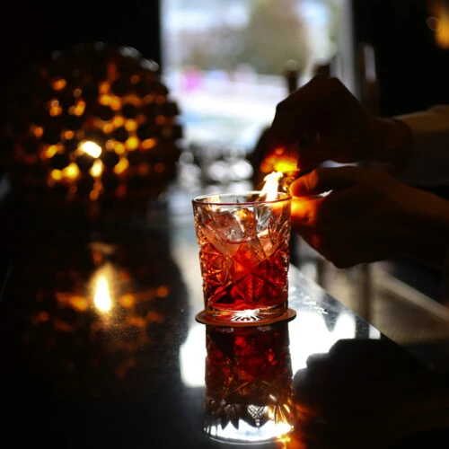 A bartender ignites citrus oils over a classic cocktail, creating a dramatic flame in a dimly lit bar.