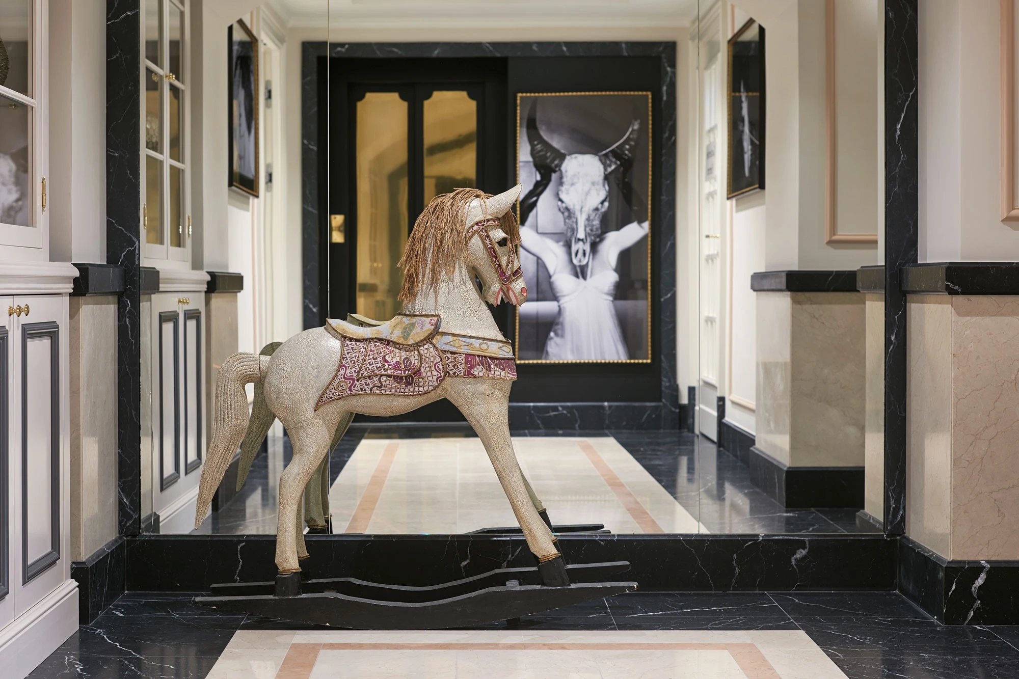 A decorative rocking horse stands in a stylish hallway with marble floors and modern artwork.