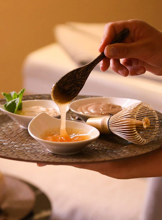 A traditional bamboo whisk lies next to a dish of creamy spa treatment, surrounded by coffee beans and elegant ceramic bowls.