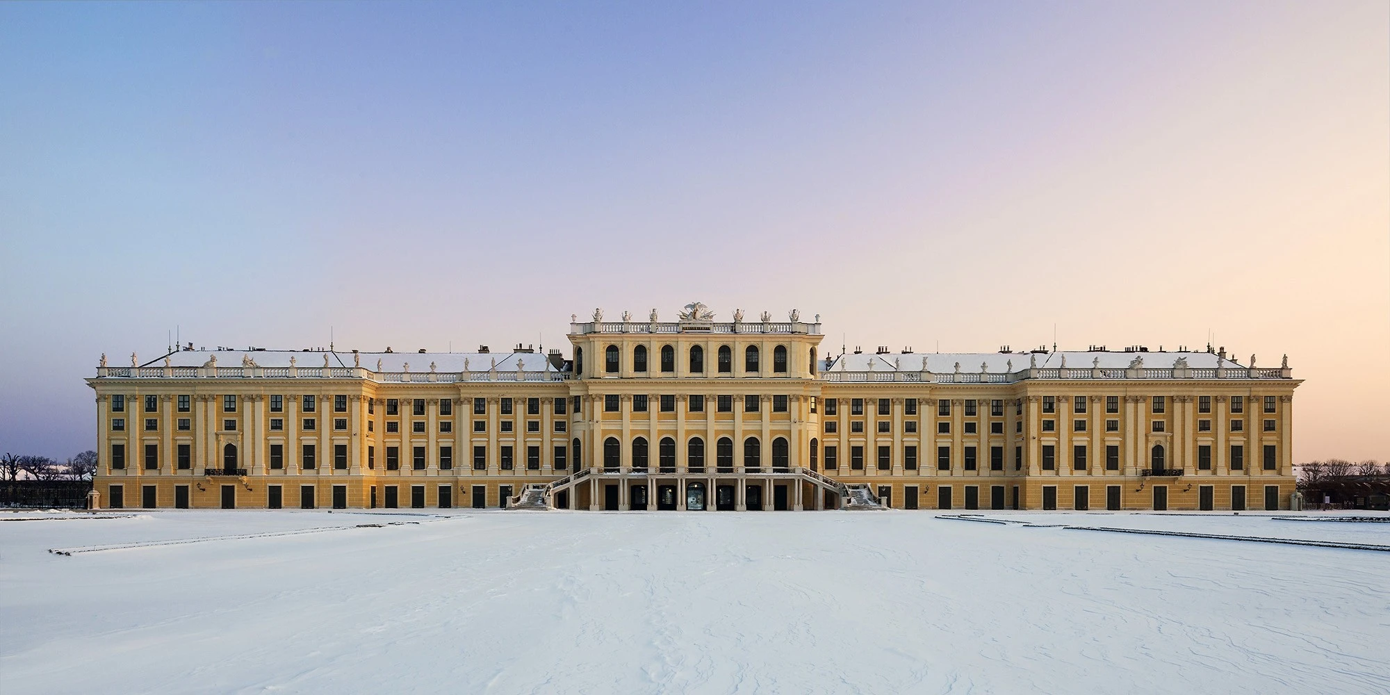 Schönbrunn Palace stands serene under a soft winter twilight, its yellow façade contrasting with the blanket of snow.