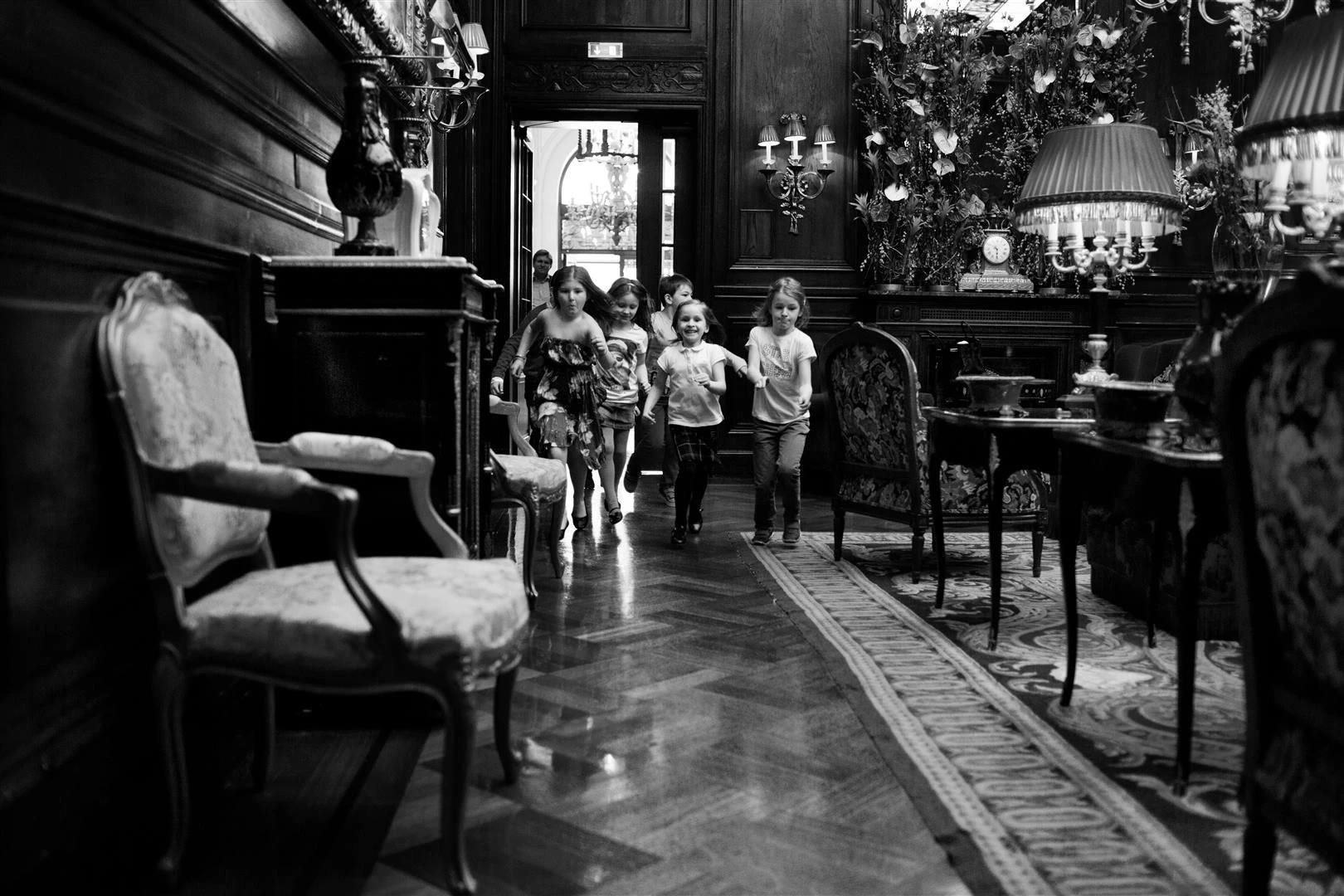 A group of children are running joyfully through an opulent, classic-style hotel hallway.