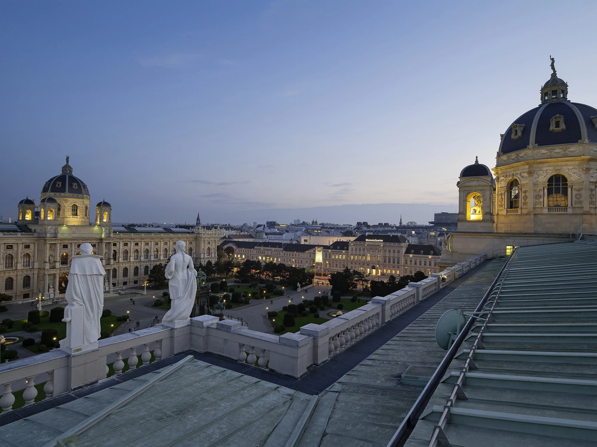 The image captures a twilight view over Vienna’s Maria-Theresien-Platz, featuring the symmetrical domes of the Kunsthistorisches and Naturhistorisches Museums, with statues overlooking the illuminated square.