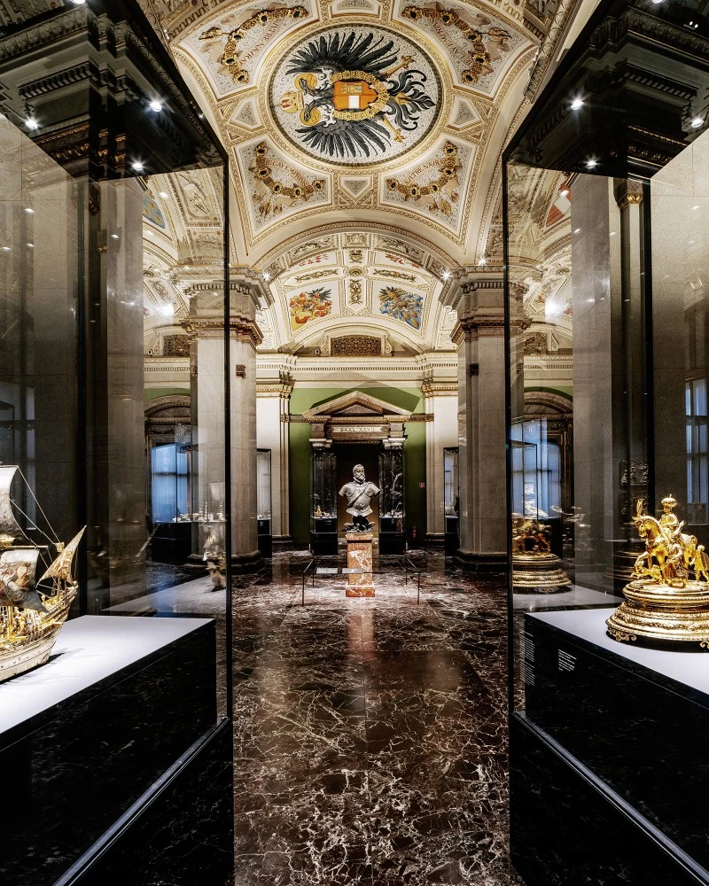 The image shows a richly decorated gallery in Vienna’s Kunsthistorisches Museum, with marble floors, gilded artifacts, and a grand ceiling featuring the imperial double-headed eagle.