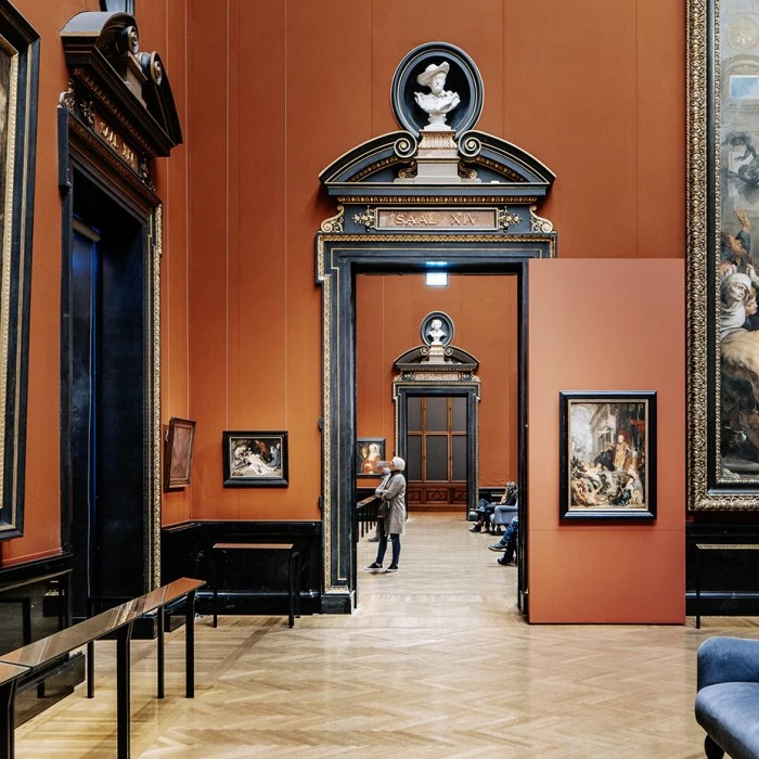 The image shows a grand gallery hall in the Kunsthistorisches Museum in Vienna, adorned with large Baroque paintings, ornate frames, and deep red walls.