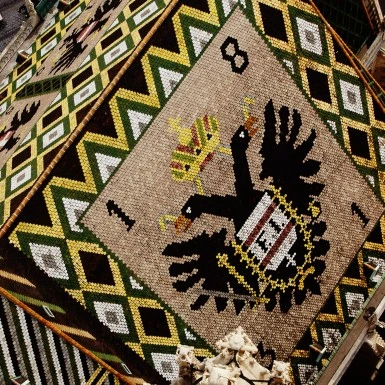The image shows the colorful tiled roof of St. Stephen’s Cathedral in Vienna, featuring the Austrian coat of arms with a double-headed eagle in intricate mosaic patterns.