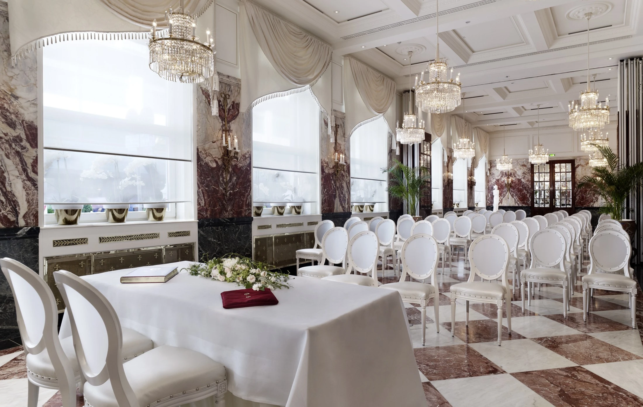 Rows of white chairs face a flower-adorned table, set for a wedding in an elegant, chandelier-lit marble hall.
