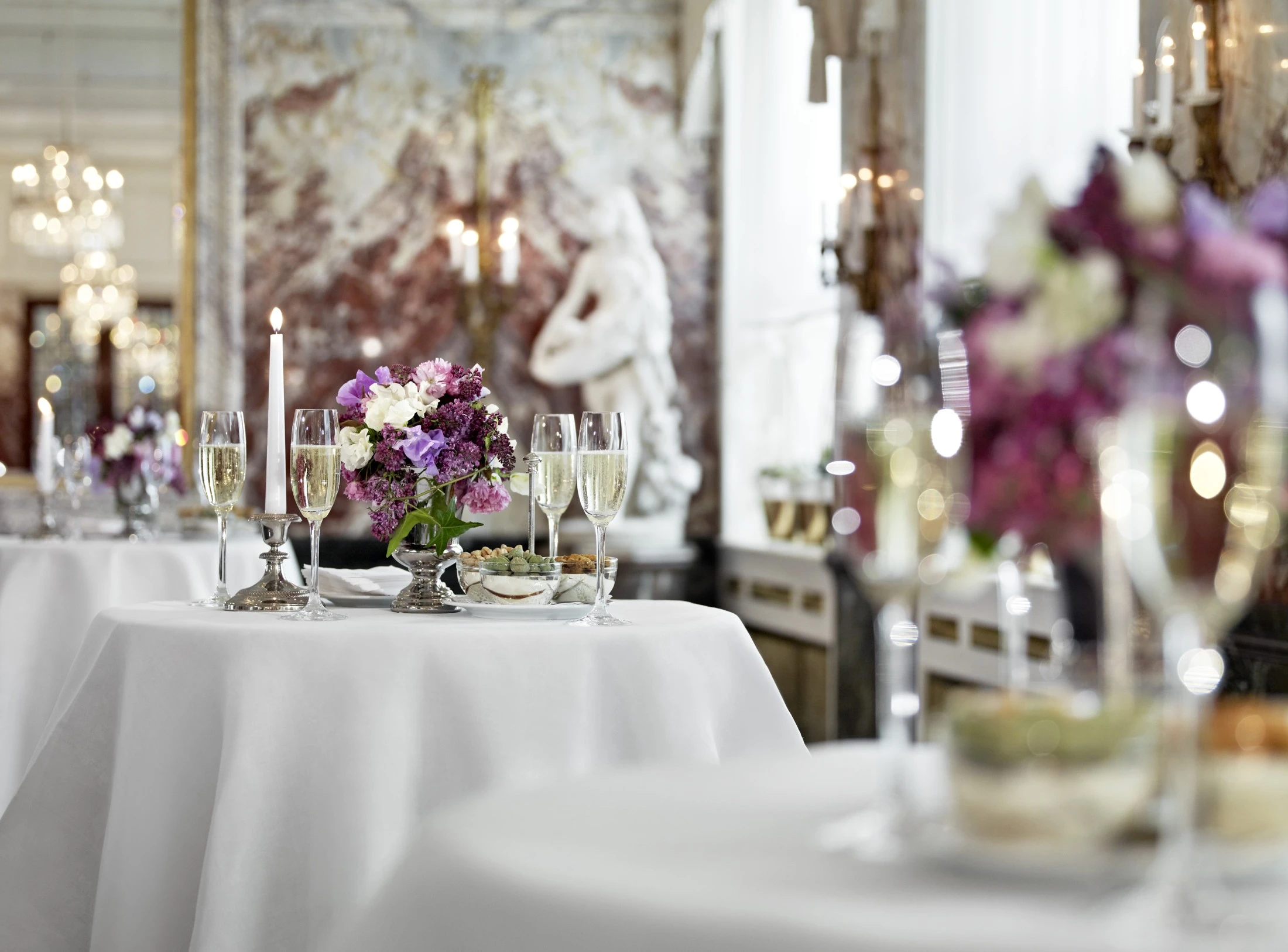 A table set with champagne glasses, flowers, and snacks stands elegantly in a marble hall.
