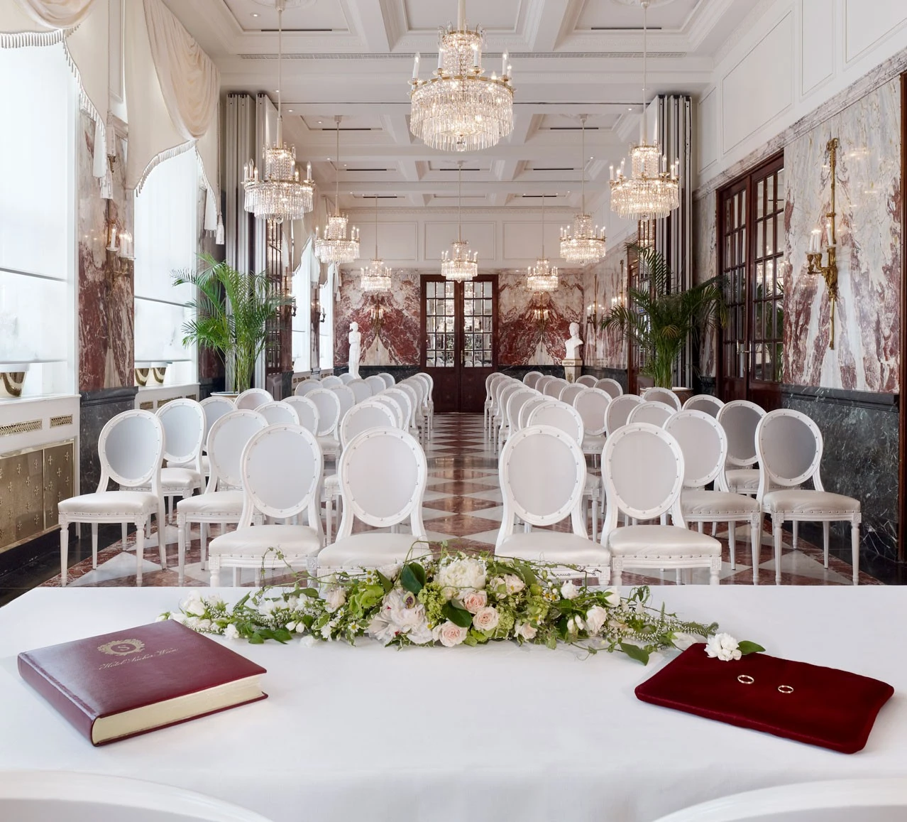 Rows of white chairs face a flower-adorned table, set for a wedding in an elegant, chandelier-lit marble hall.