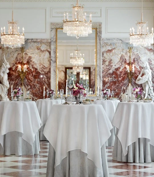 Tall cocktail tables with white linens stand elegantly under chandeliers in a grand marble hall.