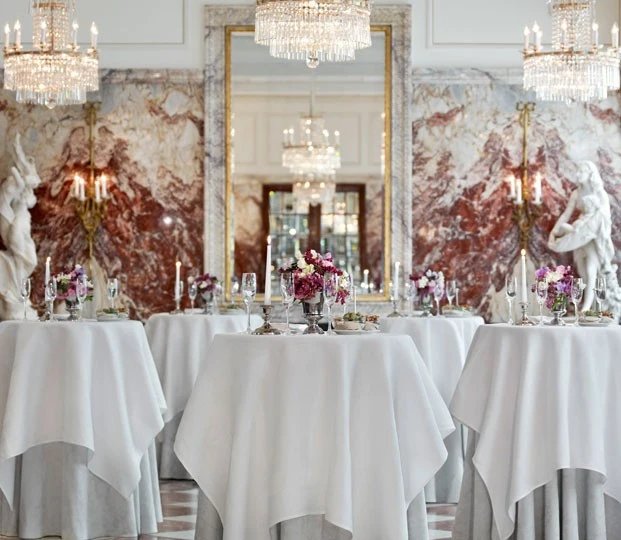 Tall cocktail tables with white linens stand elegantly under chandeliers in a grand marble hall.