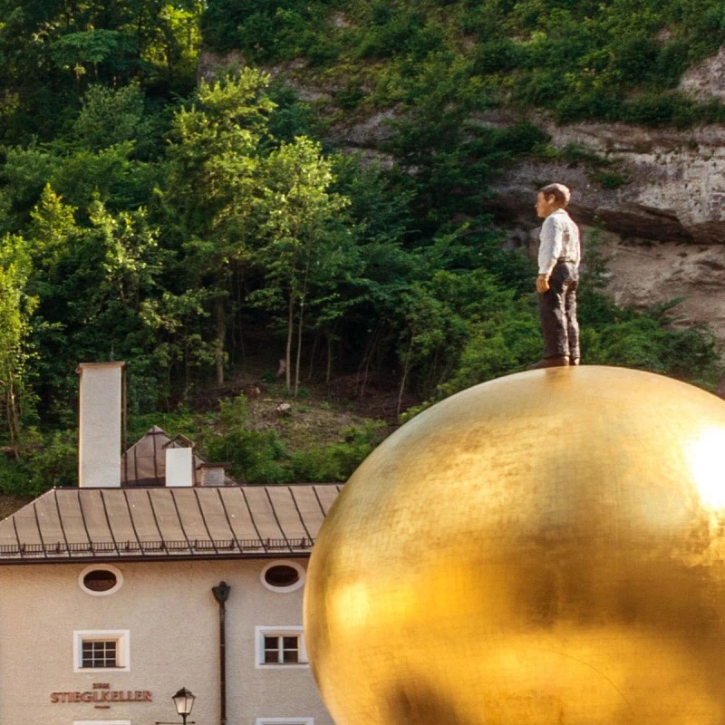 This image shows the modern art sculpture *Sphaera* in Salzburg, featuring a man standing atop a giant golden sphere.