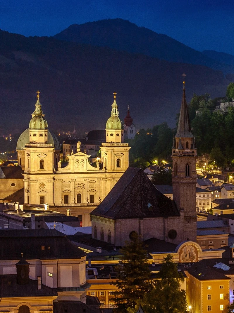 This stunning nighttime view of Salzburg showcases the illuminated DomQuartier with its twin domes and spires glowing against the dark silhouette of the surrounding mountains.
