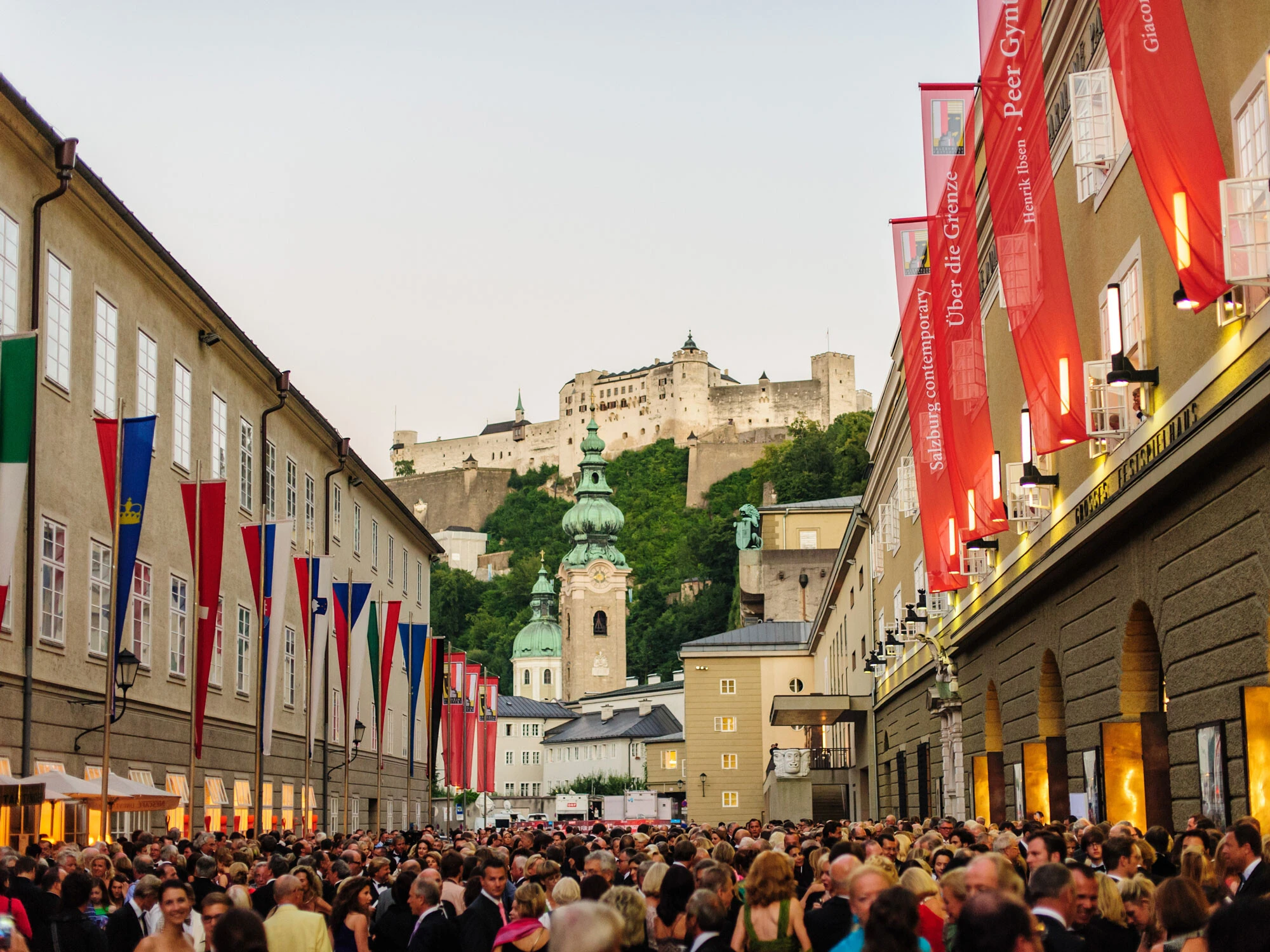 The image shows Salzburg’s historic skyline with Hohensalzburg Fortress towering above church domes and colorful festival flags.