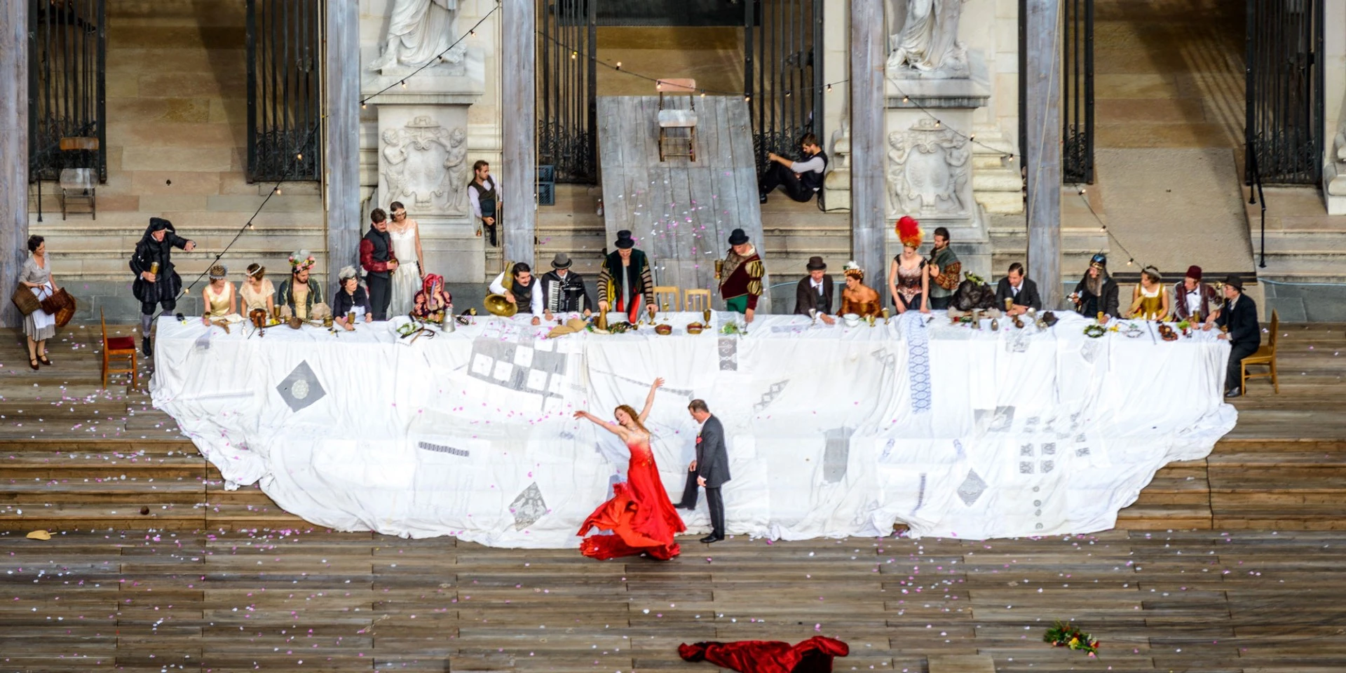 A dramatic outdoor performance at the Salzburg Festival features costumed actors, musicians, and a dancer in red.
